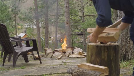 a man chops firewood near an outdoor fire pit surrounded by chairs and trees