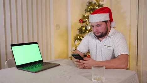 Hombre-Enfocado-Concentrado-Sentado-En-Una-Mesa-Desplaza-Su-Teléfono,-Mira-A-La-Cámara-Y-A-La-Computadora-Portátil-Con-Pantalla-Verde,-Usando-Sombrero-De-Santa