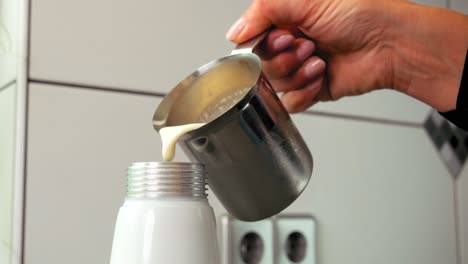 disparo estático en cámara lenta de una jarra de leche de hierro sostenida por la mano de una mujer mientras vierte crema fresca en un dispensador de crema para crema batida para hornear