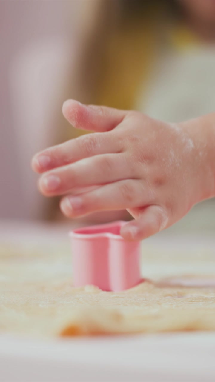 Jovem garota usa um cortador de biscoitos rosa para pressionar formas em massa estendida, criando recortes em forma de coração. As mãos dela se movem firmemente sobre uma superfície de cor clara enquanto a garota completa cada impressão.