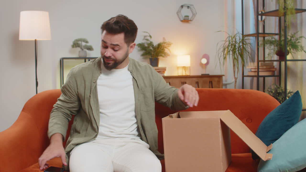 hombre infeliz desempaquetando una caja de cartón sintiéndose insatisfecho por conseguir auriculares inalámbricos dañados en casa