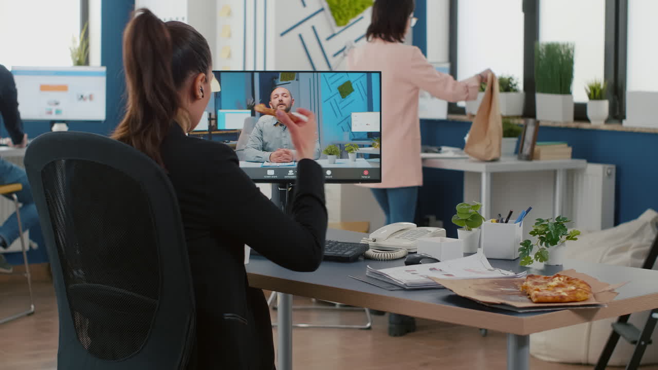 Direktør spiser et pizzastykke og diskuterer med en fjernkollega under et online videomøde.
