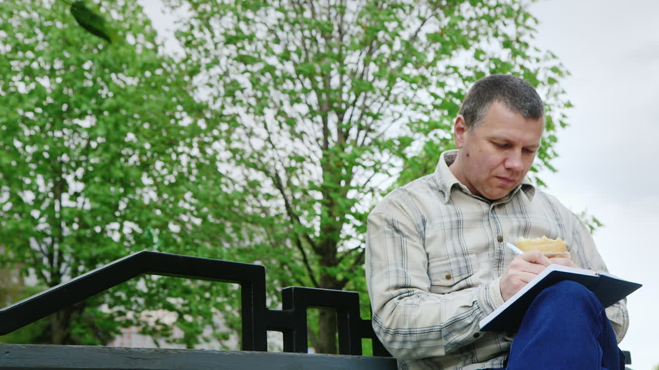 공원 벤치에서 패스트푸드를 먹는 남자는 공책에 무언가를 씁니다.