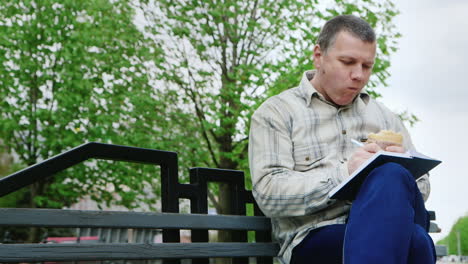 man eating fast food on a park bench writes something in a notebook