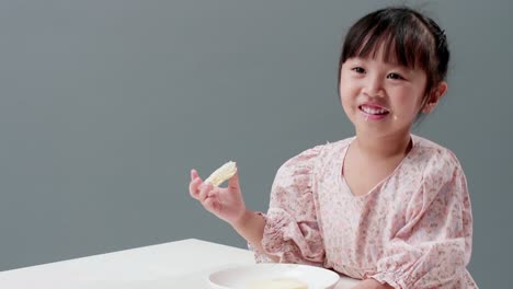 행복한 작은 아시아 소녀는 눈을 문지르고 미소를 지으며 집에서 바삭바삭한 버터 토스트를 먹습니다.