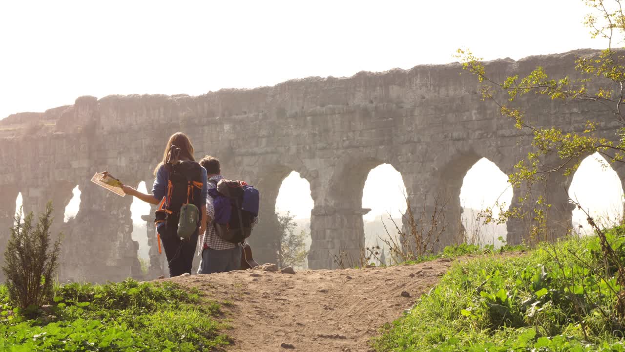 joven pareja encantadora mochileros turistas caminando de la mano hacia el acueducto romano arcos en el parco degli aquedotti ruinas del parque en roma en romántico amanecer brumoso con guitarra y saco de dormir cámara lenta