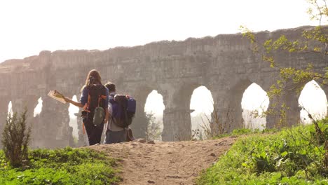 young lovely couple backpackers tourists walking holding hands toward roman aqueduct arches in parco degli acquedotti park ruins in rome on romantic misty sunrise with guitar and sleeping bag slow motion