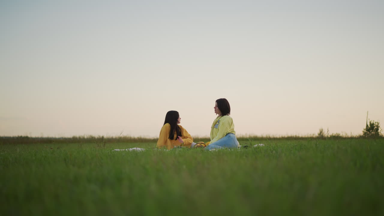 두 여성이 웃고 있는 황금 시간대의 목초지 피크닉, 담요 위에서 여행 친구들이 이야기를 나누고, 생생한 초록색 풀과 부드러운 황혼 하늘, 장난기 넘치는 몸짓과 수평선 너머로 따뜻하게 빛나는 광채