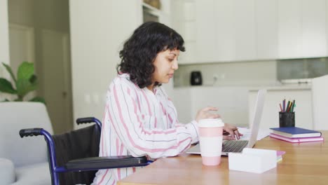 Thoughtful-biracial-disabled-woman-in-wheelchair-using-laptop-in-living-room-and-drinking-coffee