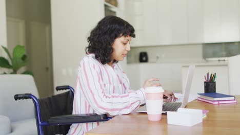 mujer discapacitada biracial pensativa en silla de ruedas usando una computadora portátil en la sala de estar y bebiendo café
