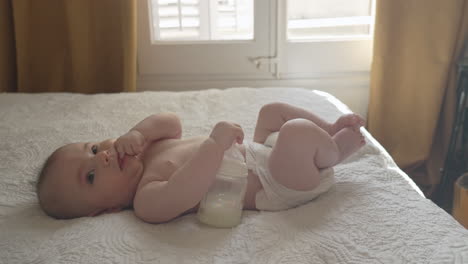 bebé contento acostado boca arriba sosteniendo su botella de leche al lado en un dormitorio blanco y acogedor