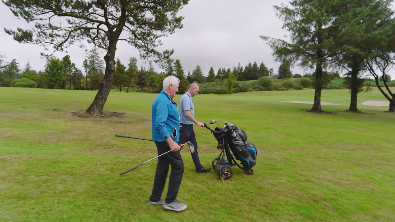 Male golf players walking with clubs and talking on golf course on cloudy day