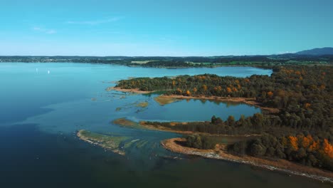 Lake-Chiemsee-in-Bavaria,-Germany-with-the-river-delta-Tiroler-Ache-in-autumn-with-blue-water-and-sky,-trees-on-an-island-and-horizon-with-the-German-and-Austrian-alps-in-the-background