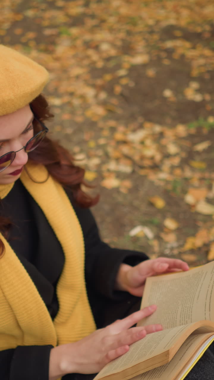 vista aérea de una mujer con el cabello rojo ondulado con boina amarilla y gafas de sol hojeando un libro en un entorno pacífico de otoño, rodeada de hojas caídas, inmersa en el pensamiento y la literatura