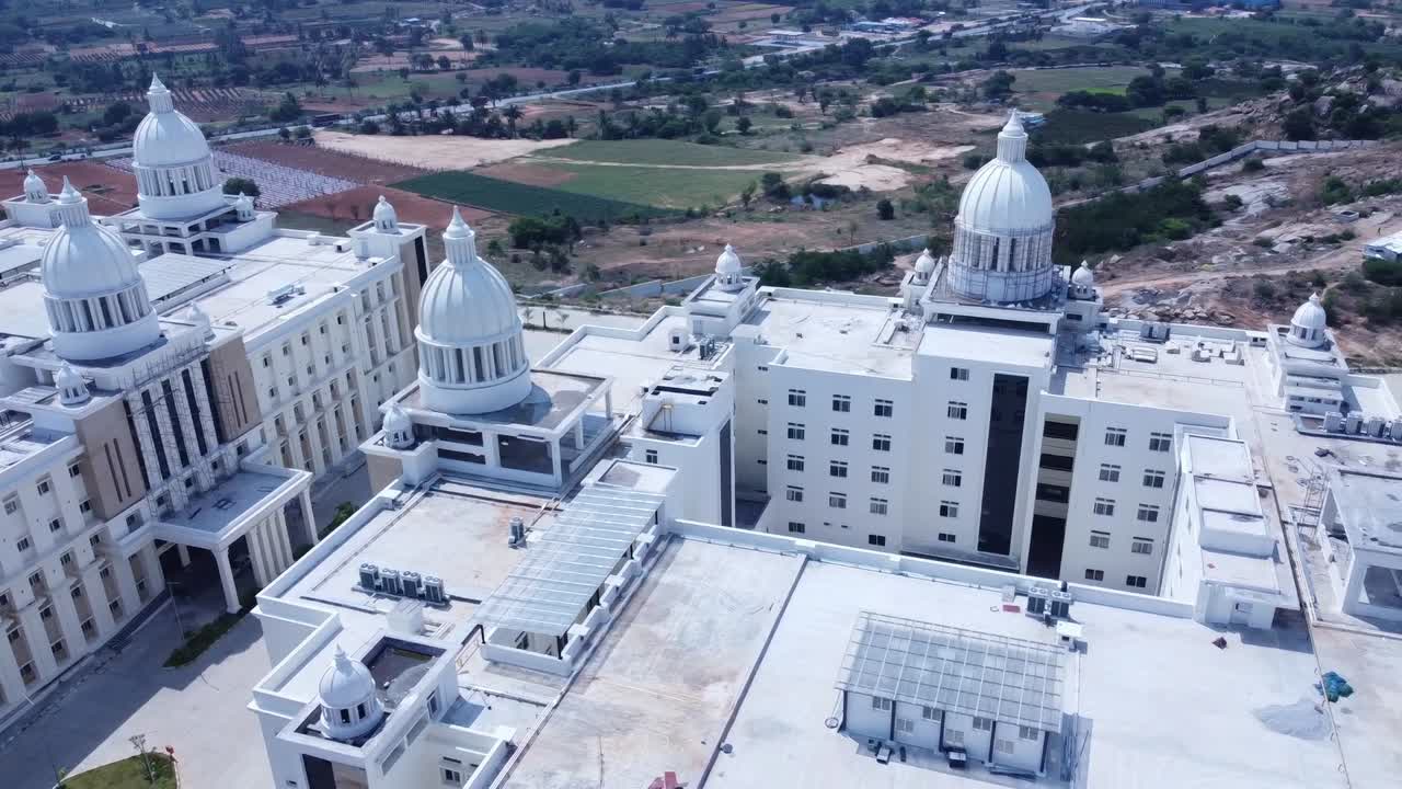 Aerial view of a grand, white institutional building with large domes. Drone pans over the massive modern architecture, revealing the surrounding rural landscape and a lake