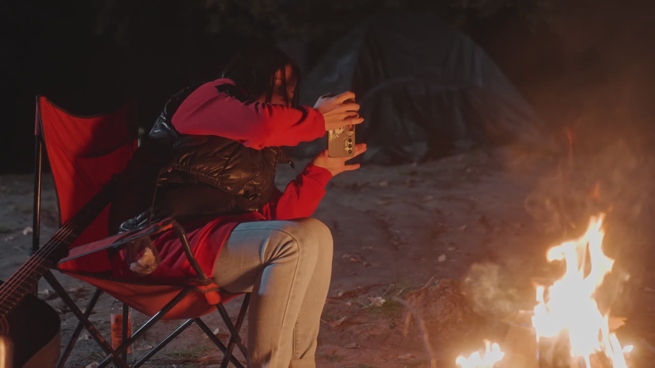 Experiência de campismo tranquila sob as estrelas com fogueira acesa e equipamento confortável. Ambiente de campismo sereno onde o indivíduo relaxa junto à fogueira crepitante, vestindo ganga e casaco colorido