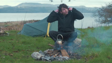 young man sitting by fire camping on island puts up hood in rain