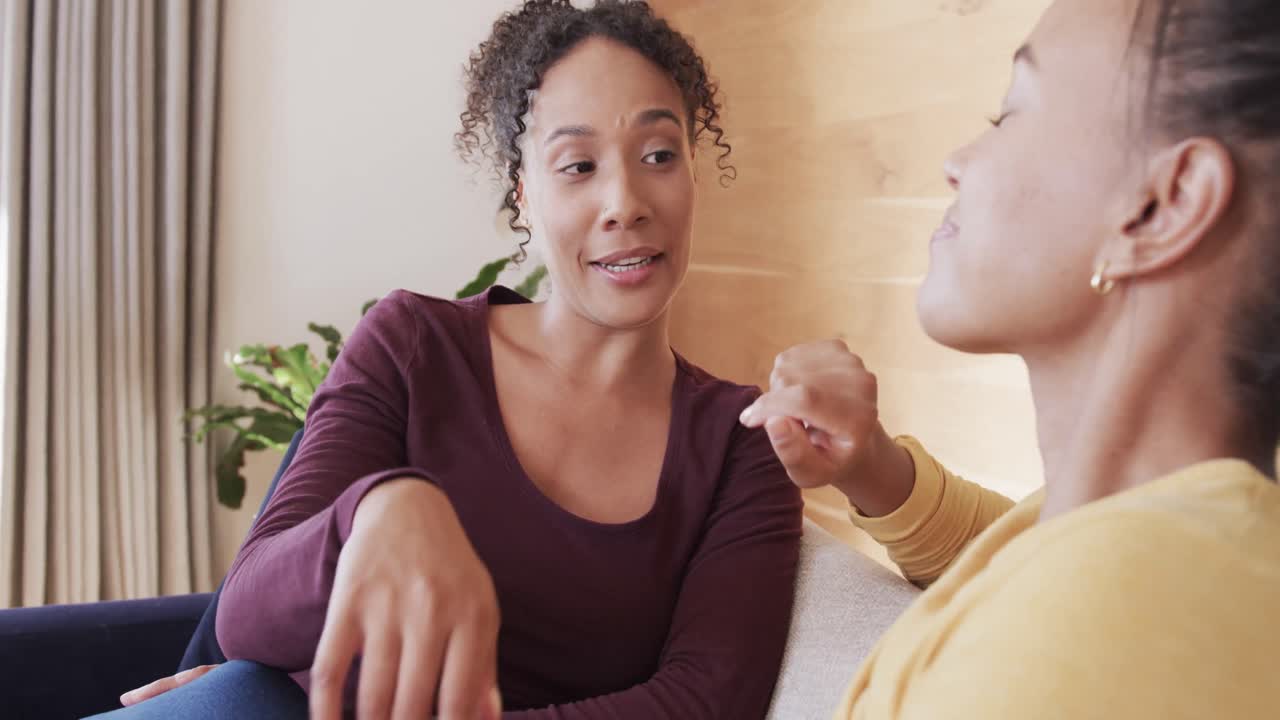 Happy biracial lesbian couple relaxing on couch together, holding hands and talking, in slow motion