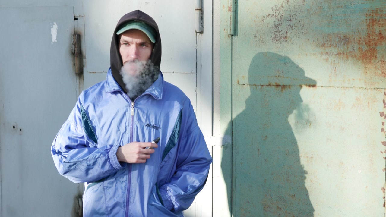 Hombre con chaqueta con capucha exhalando humo contra una puerta de metal oxidada, sombra proyectada sobre superficie desgastada, aliento frío