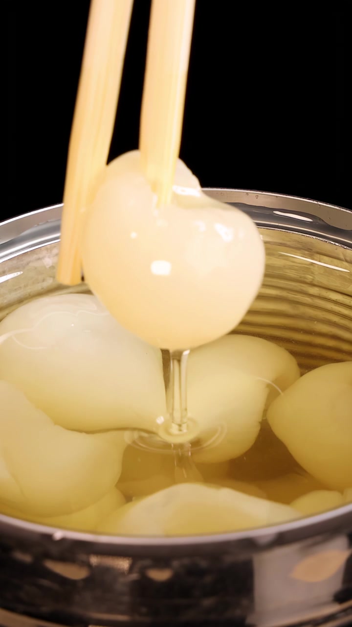 Chopsticks grasp a peeled longan fruit from syrup inside a metal can under bright, even lighting, with a close-up, steady camera view and black background