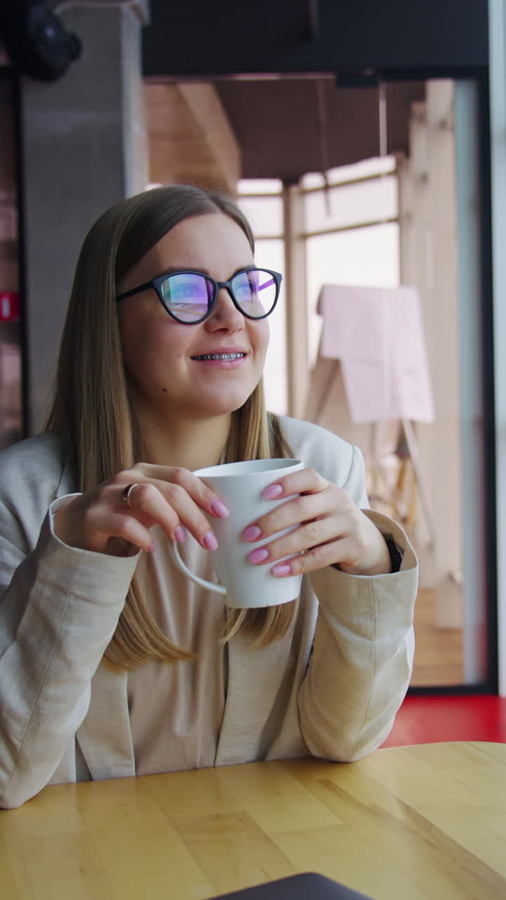 Relaxed lady drinks tea enjoying lunch time rest. Young smiling woman sitting at the table and looking in the window. Vertical video