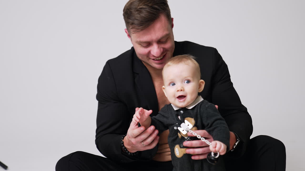 Happy Caucasian man in black jacket sitting on the studio floor with his baby. Little child trying to walk himself. White backdrop.