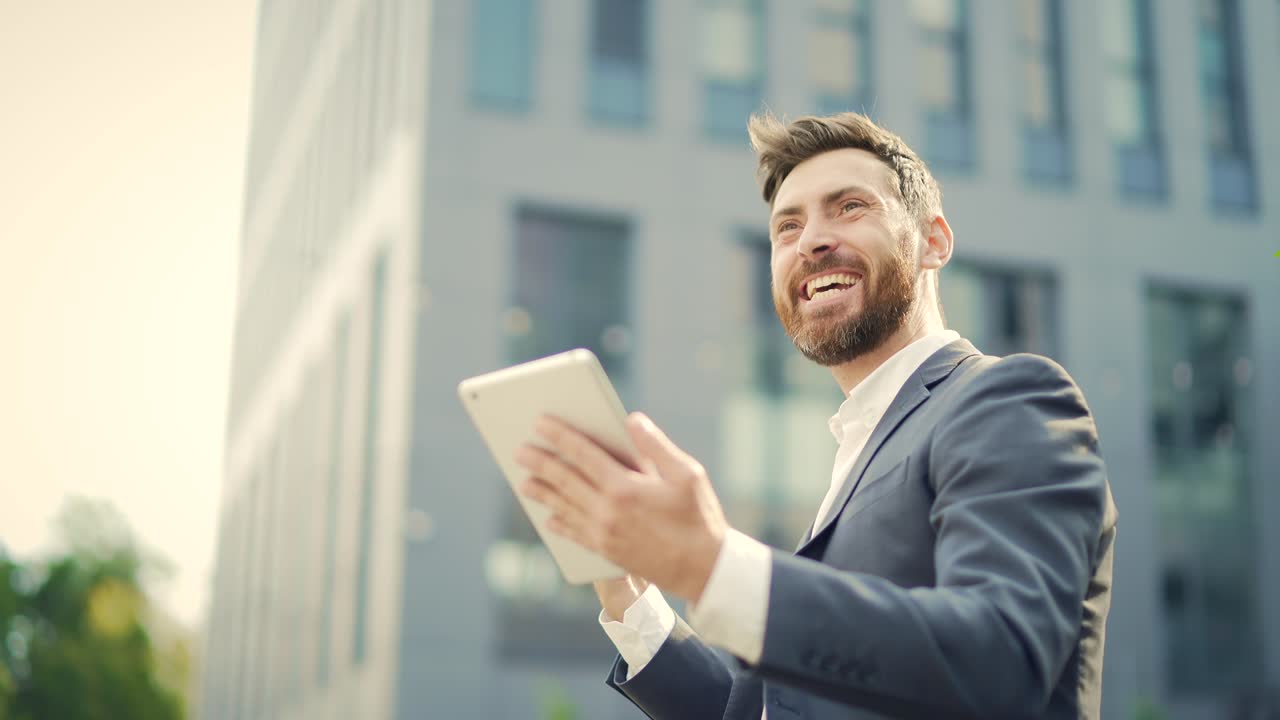 Empresario emocionado celebrando un logro, mirando su tableta digital y teléfono inteligente al aire libre. Un hombre de negocios exitoso celebra su victoria con el teléfono móvil en el exterior