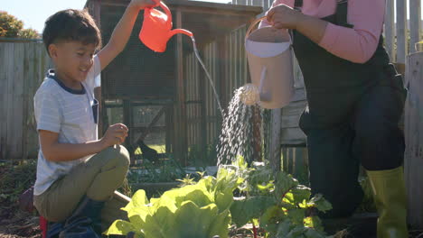 Abuela-Birracial-Mayor-Y-Nieto-Regando-Plantas-En-Un-Jardín-Soleado,-Cámara-Lenta