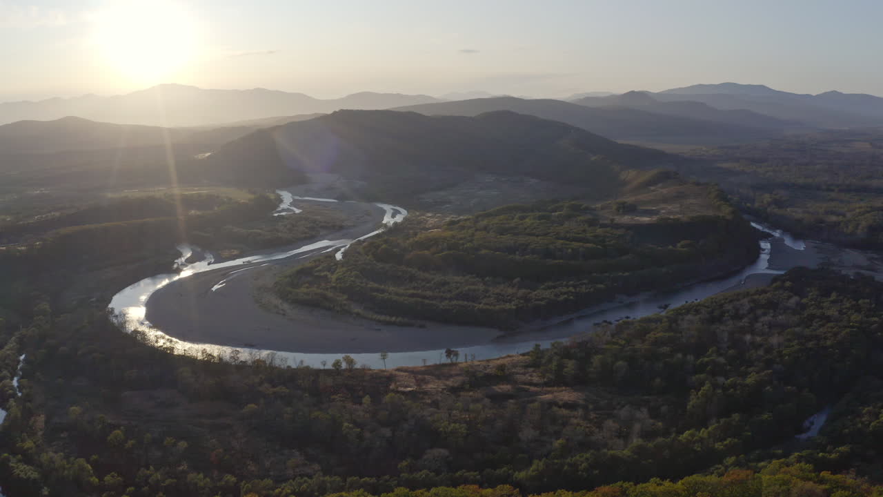 Bird's eye view of a valley with riverbed taking a one hundred eighty degree turn, the sun touching the top of mountain ridge in the background. Sunset. Aerial jib up shot.