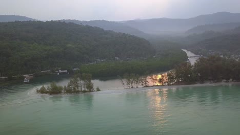 vista aérea matutina desde un dron en lo alto del cielo moviendo lentamente la cámara hacia arriba revelando el paisaje: playa de arena, costa y montañas a lo lejos también amanecer reflejado en el océano
