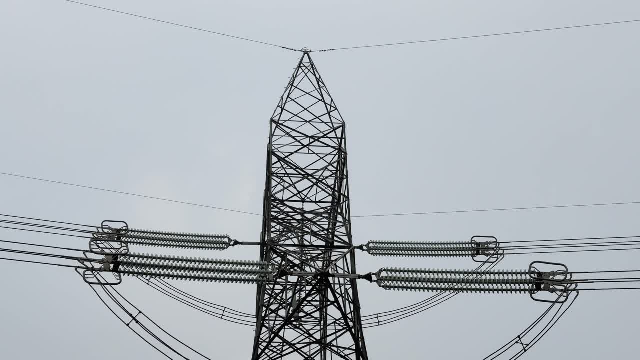 Tilt-down shot revealing the overhead ground wire mounted at the top of a high-voltage transmission tower, with steel lattice structure, insulators, and power lines visible against a neutral sky