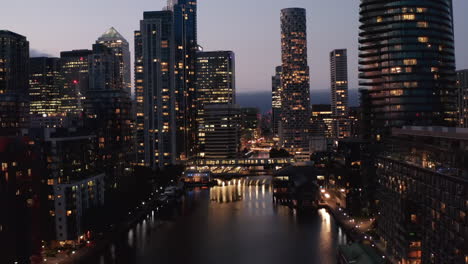 Backwards-reveal-of-skyscrapers-along-water-channel-in-Canary-Wharf-business-centre-against-twilight-sky.-City-lights-reflecting-on-water-surface.--London,-UK