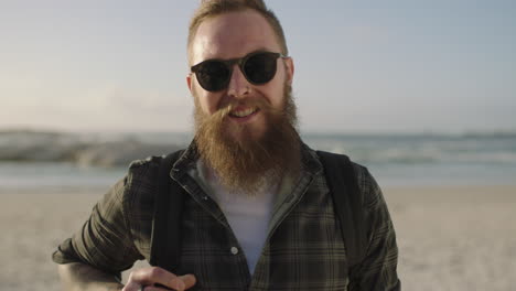 Bärtiger-Hipster-Mann-Mit-Tätowierungen-Posiert-Selbstbewusst-Am-Strand-Und-Trägt-Eine-Sonnenbrille,-Die-Glücklich-Lächelt