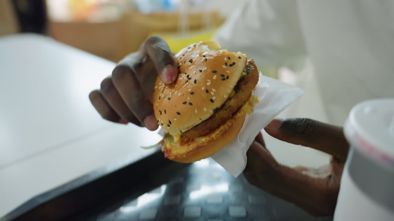 Close up of hungry man wearing white shirt holding sesame burger with two hands wrapped in napkin as he prepares to eat while seated indoors with colorful bokeh background at food court table