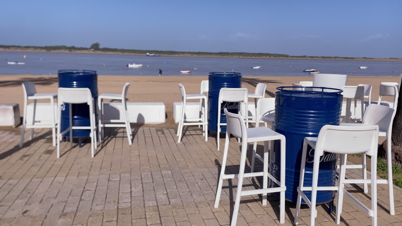 Tavoli da bar all'aperto realizzati con barili blu e sgabelli bianchi che invitano al relax in riva al fiume, con barche che riposano sull'acqua.
