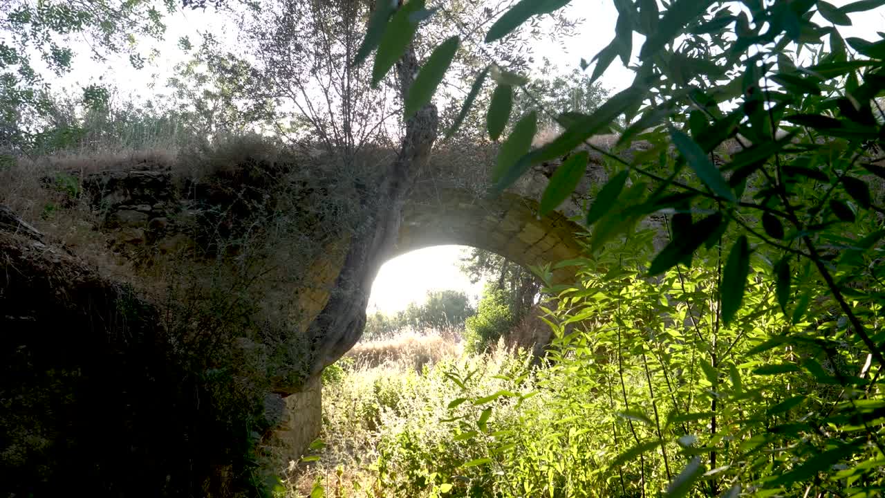Este local apresenta uma antiga ponte de pedra escondida em meio a uma vegetação exuberante. A luz quente do pôr do sol destaca a beleza do arco e das plantas ao redor.