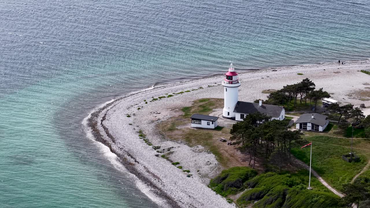 Aerial drone footage of Sletterhage Lighthouse on the coast of Denmark, showing the white lighthouse, curved shoreline, and turquoise water under bright daylight