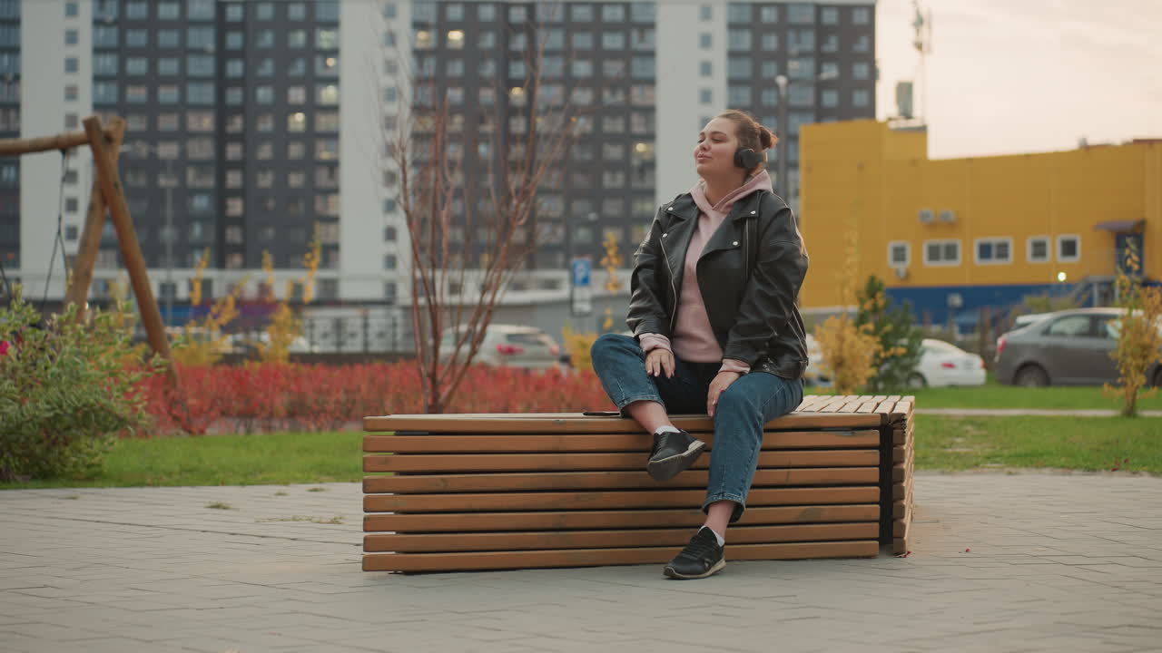 Young girl seated on wooden bench outdoors wearing black leather jacket listens to music through headphones smiling and dancing slightly as cars park and office buildings in background