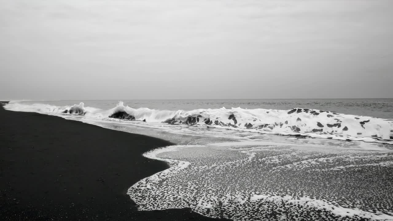 Black and white video still of waves crashing on a dark sandy beach, captured from a low angle