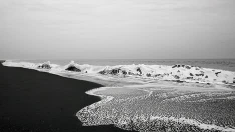 black and white video still of waves crashing on a dark sandy beach, captured from a low angle