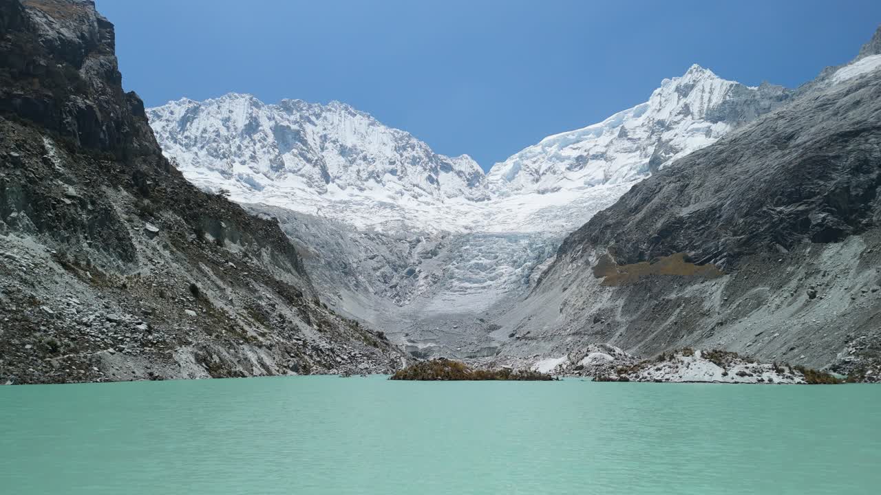 An aerial flyover begins above Laguna Llaca’s stunning turquoise waters and tilts up, revealing dramatic rocky slopes and snow-capped Andean mountains in Peru’s Cordillera Blanca