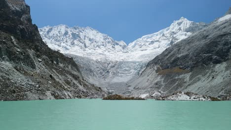 an aerial flyover begins above laguna llaca’s stunning turquoise waters and tilts up, revealing dramatic rocky slopes and snow-capped andean mountains in peru’s cordillera blanca