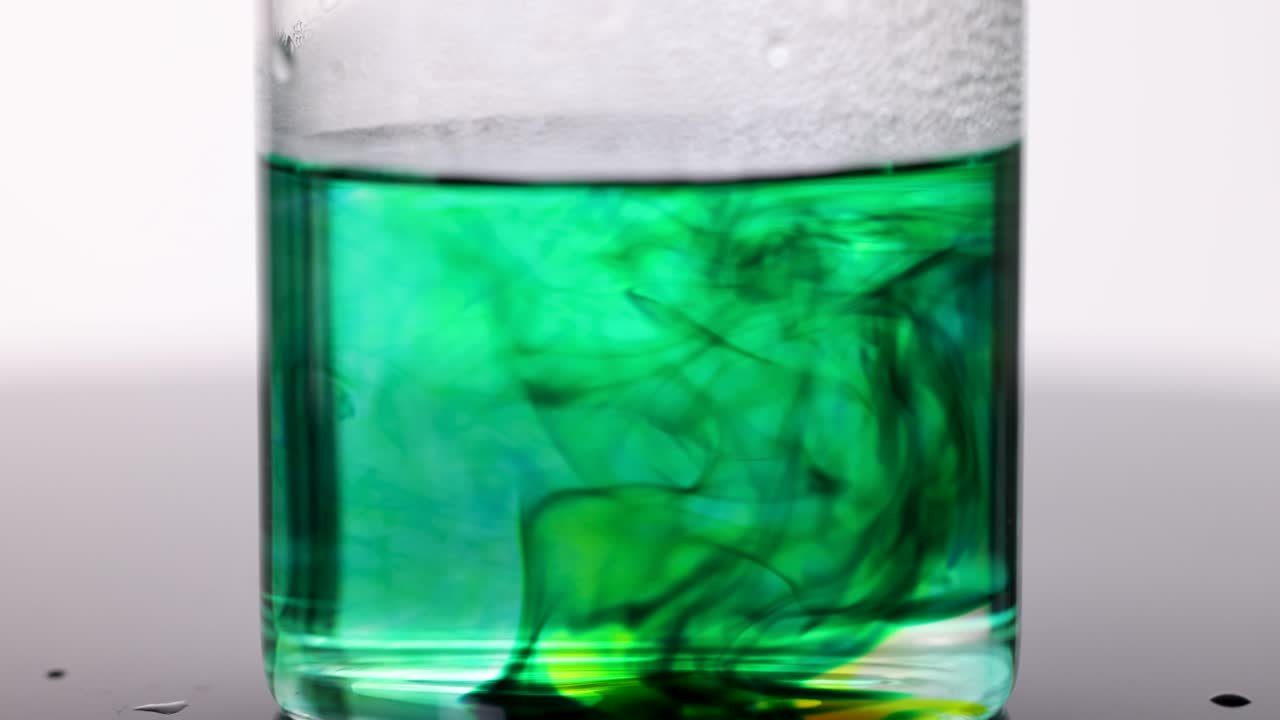 Green food coloring swirls and disperses in a clear glass of hot water, studio lighting