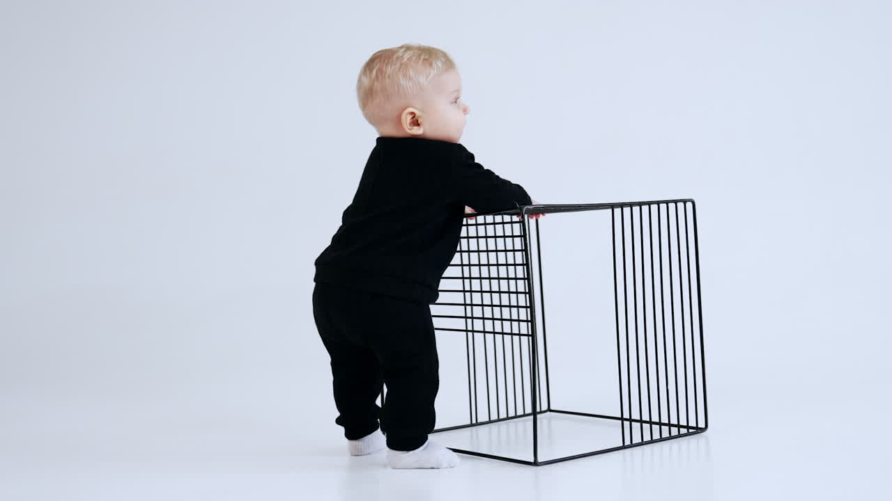 Blond Caucasian baby boy wearing black suit stands in studio. Little kid taps his hand by the grate cube. White backdrop.