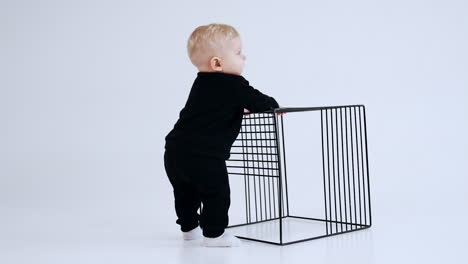 blond caucasian baby boy wearing black suit stands in studio. little kid taps his hand by the grate cube. white backdrop.