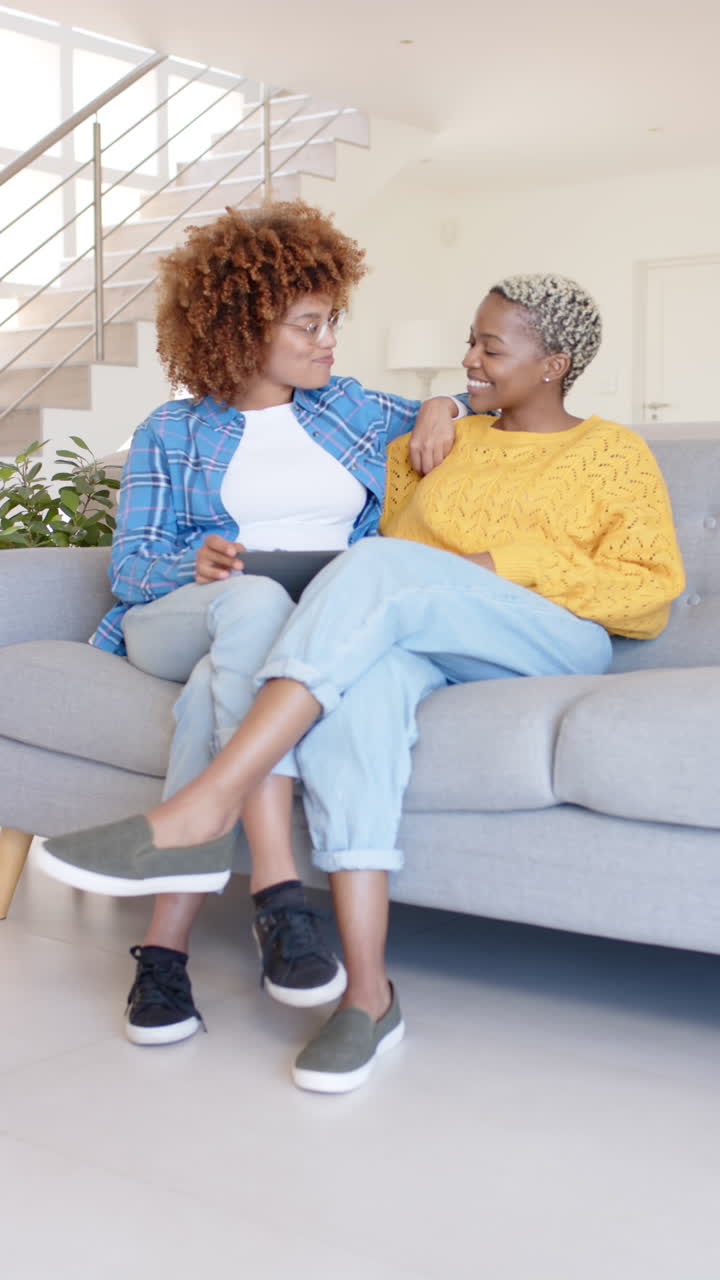 Vertical video of happy diverse female couple using tablet and talking in slow motion
