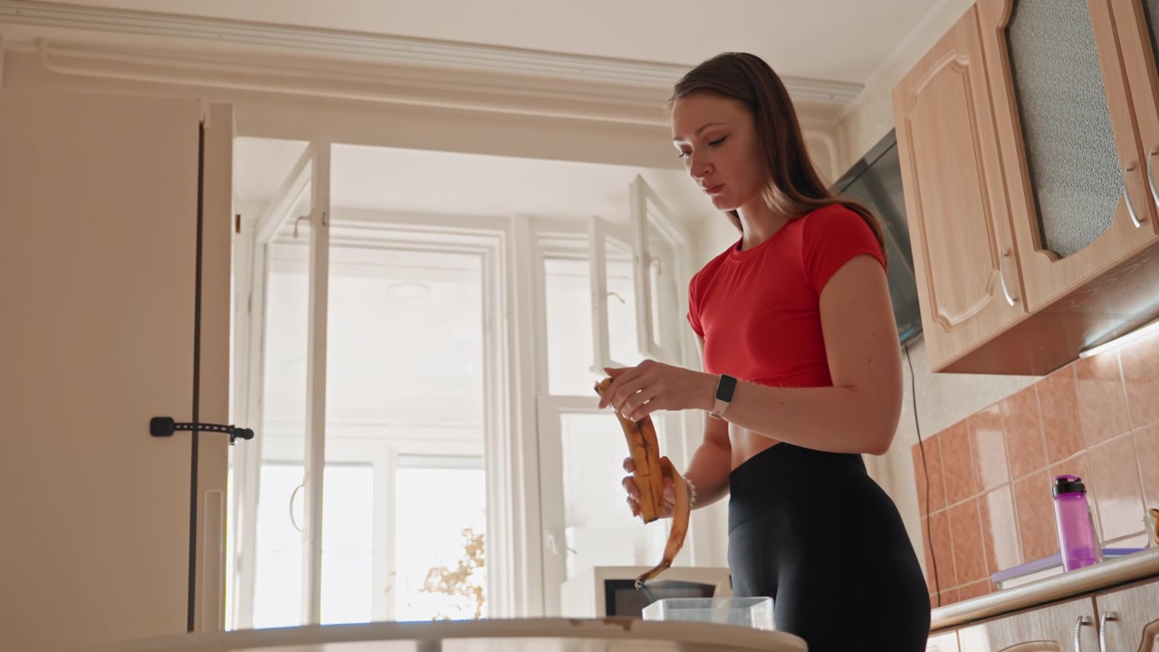 Giovane donna in cucina che apre il pane, fornaia casalinga che prepara lo spuntino mattutino con la calda luce del sole, in piedi al bancone mentre affetta il pane, con una maglietta rossa casual, paraschizzi moderno piastrellato, lavello e bottiglia sul bancone, in un'atmosfera calma