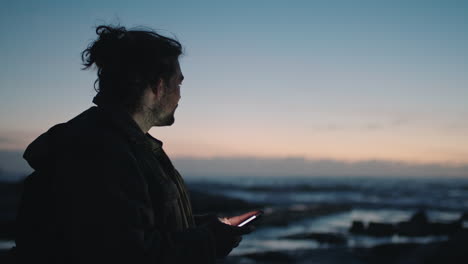 young man texting on phone near sea at sunrise sunset