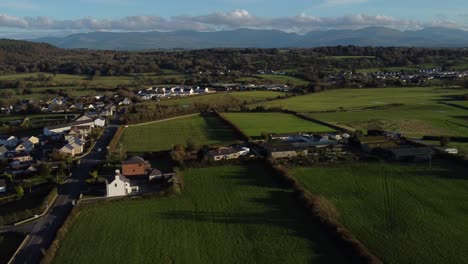 Aerial-view-panning-across-small-town-Welsh-community-farmland-countryside-with-Snowdonia-mountain-range-on-the-horizon