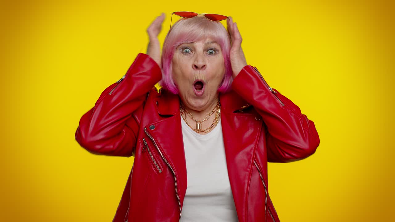 흥분한 시니어 여성이 놀라서 손을 들어 카메라를 쳐다보며 갑작스러운 승리에 충격을 받았습니다.